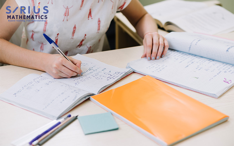 Person writing math equations in a squared notebook at a desk with study materials and open books.