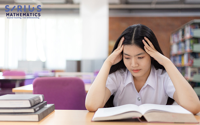 Student reading at a desk in a quiet library with bookshelves, purple chairs, and stacked books.